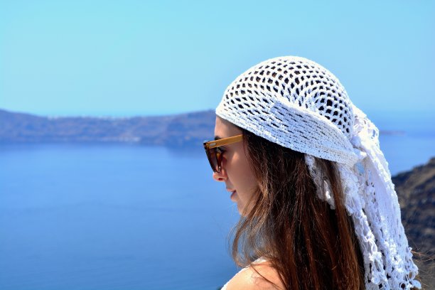 年轻的黑发女子的肖像，戴着太阳镜和波西米亚白色钩针帽，欣赏风景，夏天的地中海，蓝天为背景，费拉，圣托里尼岛，希腊，欧洲图片下载
