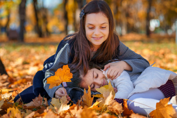 在一个秋天的城市公园里，两个女孩坐在一片黄枫叶的空地上，孩子们在玩耍和享受，在一棵树旁摘树叶，大自然美丽，阳光明媚图片下载