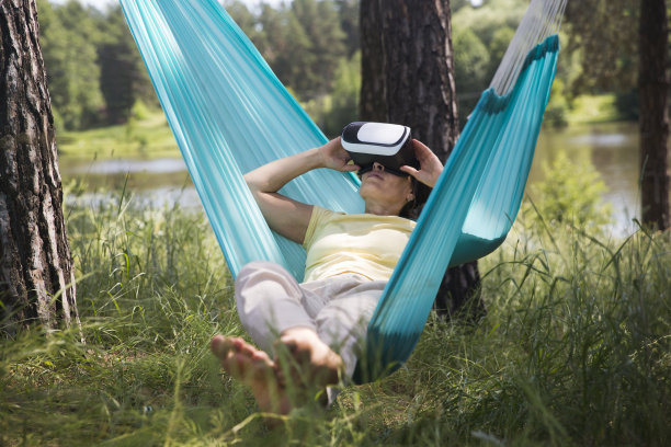 戴着虚拟现实眼镜在吊床上玩游戏的中年妇女在户外放松。副本的空间。图片下载