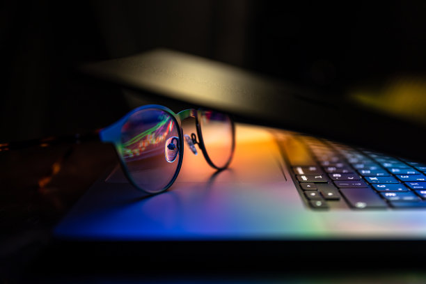一台笔记本电脑在黑暗中半闭着，带着彩色的光芒和眼镜。图片下载