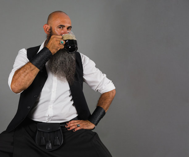 一个剃了光头、留着长胡子的成熟男人，一边喝着黑啤酒，一边看着灰色背景下的镜头图片下载