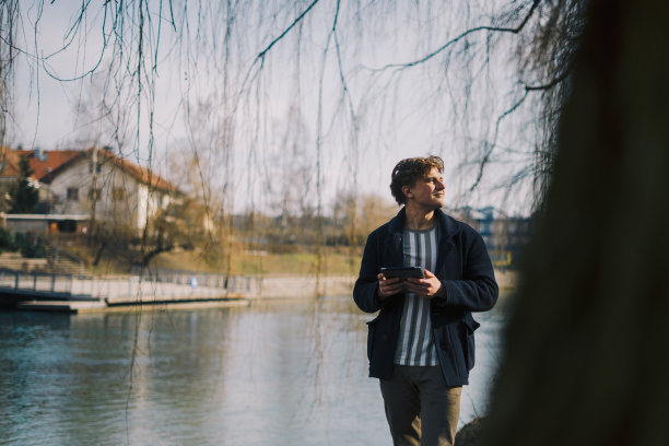 一个年轻人在河边的公园外使用数码平板电脑的正面照片。副本的空间。库存图片图片下载