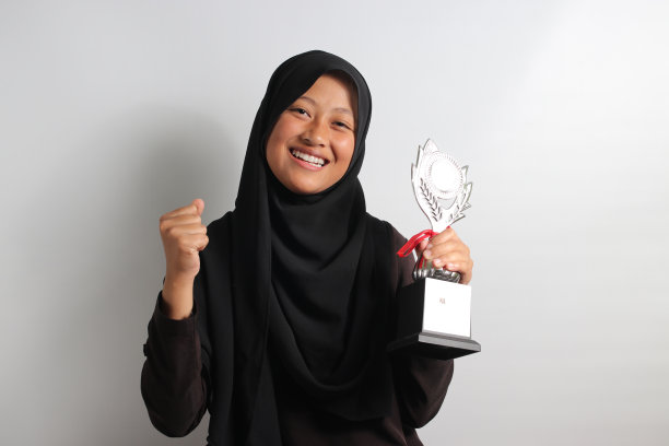 聪明的年轻亚洲女学生戴着头巾，在白色的背景上举起奖杯。图片下载