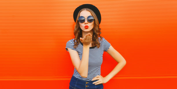 美丽的年轻女子在橙色背景上戴着黑色圆帽，吹着她的嘴唇，送出甜蜜的飞吻图片下载