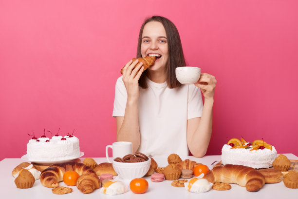 快乐满意的棕色头发的女人，穿着白色t恤，坐在桌子旁，吃着自制的糕点，咬着羊角面包，端着茶杯，吃着甜点，在粉红色的背景下快乐地孤独着。图片下载