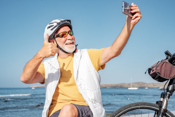 视频通话概念。面带微笑的骑自行车老人戴着头盔坐在沙滩上，骑着电动自行车用手机视频聊天。老年人积极向上图片下载