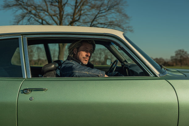 一名穿着牛仔裤夹克的金发男子坐在一辆老式美国肌肉车的副驾驶座位上，看着窗外。图片下载