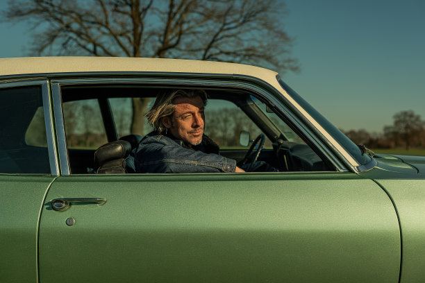 一名穿着牛仔裤夹克的金发男子坐在一辆老式美国肌肉车的副驾驶座位上，看着窗外。图片下载