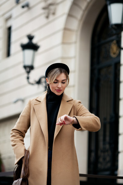 优雅的时装模特站在城市的街道上看手表。时尚的年轻女子穿着时髦的衣服配饰发带，黑色高领毛衣，米色外套图片下载