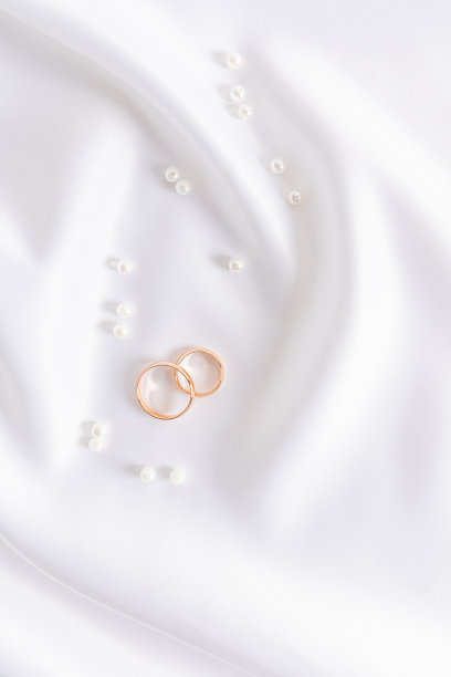 美丽精致的婚礼背景，白色优雅的缎面织物，两个金婚戒和散落的珍珠。垂直的观点。图片下载