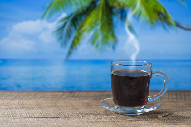 在热带海滩咖啡馆，阳光明媚的夏日里，热黑咖啡的玻璃杯上有海水、椰树和蓝天背景的木制桌子，特写，复制空间图片下载