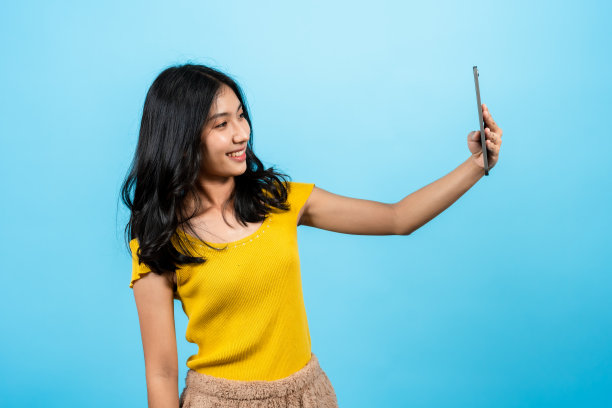 肖像，长发亚洲女孩穿着紧身t恤，站在笔记本电脑上与男友视频通话，看着男友的脸，开心地笑着，蓝色背景的室内工作室。图片下载