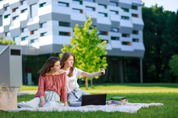 年轻的女友们坐在公园的草地上，用智能手机自拍，时髦的女孩们用手机拍一张普通的照片，发布在网络上，在大学校园里休息图片下载