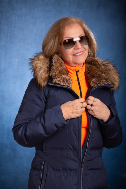 一个60多岁的女人的肖像，穿着一件冬衣，里面穿了一件亮橙色的上衣，戴着太阳镜。她微笑着把目光移开镜头图片下载