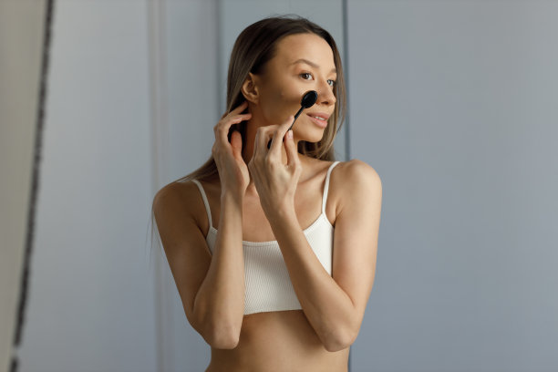 美丽的年轻微笑的金发女郎，长头发，穿着白色短上衣，在脸上的痘痘上涂抹粉底。用专业刷子涂抹浅色消光粉图片下载