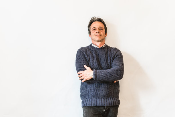 快乐的笑着的男人，微笑着的中年老人穿着蓝色的毛衣双臂交叉站在白色的墙上，肖像图片下载