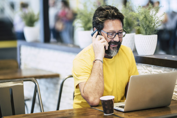 一个年轻的大胡子男人坐在咖啡馆或联合办公空间忙着打电话，用无线连接笔记本电脑免费工作。新商务人士生活方式理念图片下载