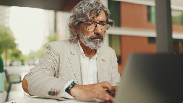 戴着眼镜留着胡子的成熟商人，坐在户外咖啡馆里用笔记本电脑做笔记。成功的男人坐在自然咖啡馆的桌子上，移动办公室自由职业者图片下载