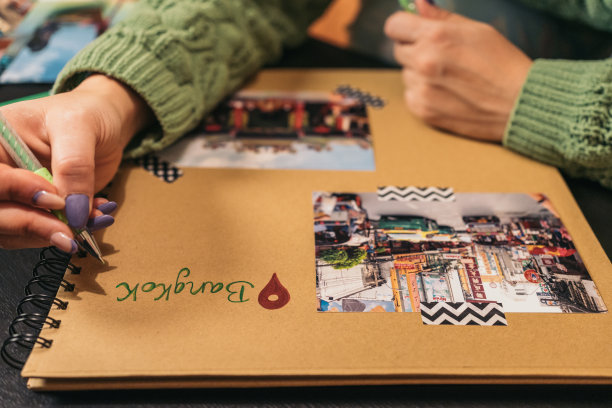 一个无法辨认的年轻女子的手在一个手工制作的牛皮纸相册上写着“曼谷”这个词，相册里有旅行照片和washi胶带。图片下载