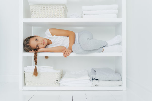 带着小辫子的宁静女孩的横照躺在白色的控制台架子上，穿着休闲的衣服，周围是叠好的亚麻衣服和一盆新鲜的衣服，在帮助母亲后休息图片下载