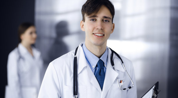 友好的年轻男医生拿着写字板站在现代诊所里。女同事在医生的后台忙着填写病历表。医学的概念图片下载