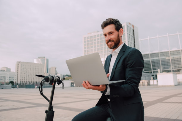 穿着正装的现代商人面带微笑，站在电动摩托车旁边，拿着笔记本电脑和工作用具图片下载