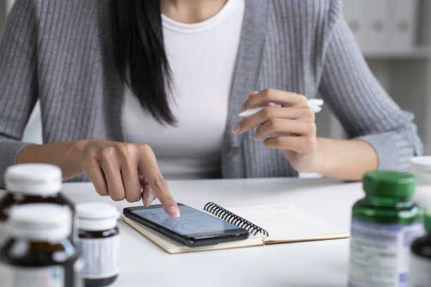亚洲女性专业营养师一边看手机一边做笔记，桌子上摆满了各种各样的膳食补充剂图片下载