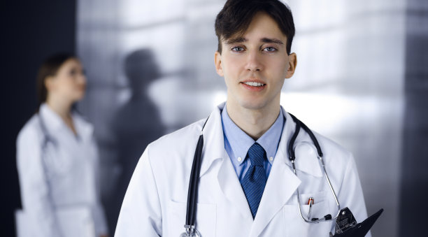 友好的年轻男医生拿着写字板站在现代诊所里。女同事在医生的后台忙着填写病历表。医学的概念图片下载