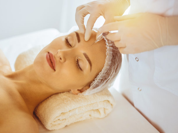 在阳光明媚的医疗中心，美女闭着眼睛接受美容注射。美容师医生用注射器给女性面部做美容手术图片下载