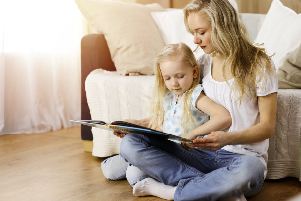 幸福的家庭。在阳光明媚的房间里，金发碧眼的年轻母亲坐在木地板上给她可爱的女儿读书。母亲的概念图片下载