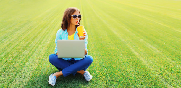 在绿色草地背景下的夏日公园里，一个女人拿着笔记本电脑喝着鲜榨果汁图片下载