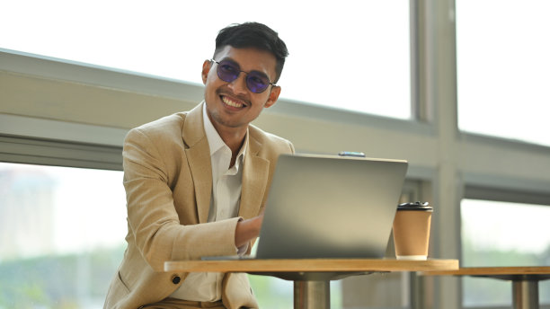 戴着光致变色眼镜，微笑的年轻商人在现代咖啡店里用笔记本电脑工作。图片下载