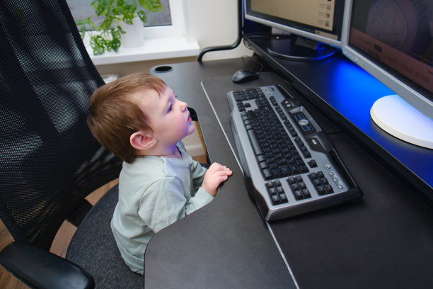 快乐的宝宝正坐在显示器旁边的电脑键盘前。一个孩子在家里的客厅里用台式电脑。两岁左右(一岁九个月)图片下载