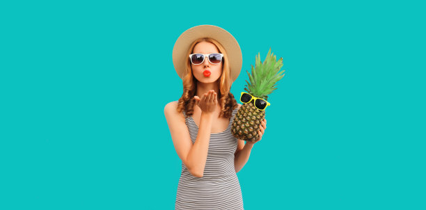 夏天，年轻女子在蓝色背景上戴着草帽和太阳镜，吹着她的嘴唇与菠萝飞吻图片下载