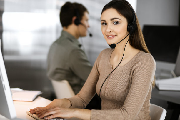 在一间现代化的办公室里，戴着耳机的年轻友好的女孩和她的同事坐在办公桌前，与公司的客户交谈。呼叫中心接线员正在工作图片下载