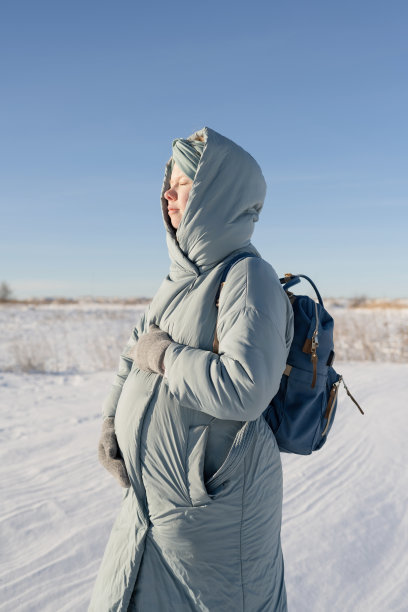 在冬天的公园里，穿着暖和的羽绒服，闭着眼睛抱着肚子，一名放松的年轻孕妇。图片下载