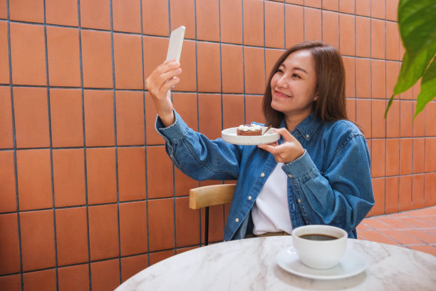 图为一名年轻女子在咖啡馆拿着蛋糕用手机自拍图片下载