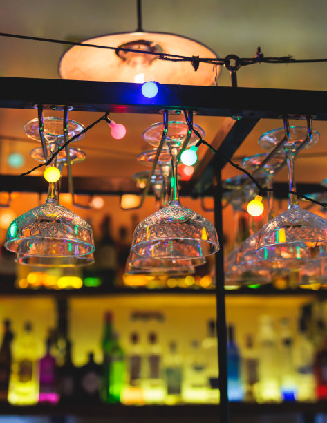 酒吧内部的细节，葡萄酒和不同的酒精鸡尾酒酒杯悬挂在吧台上，背景是酒瓶和调酒师，夜生活俱乐部有背光照明图片下载