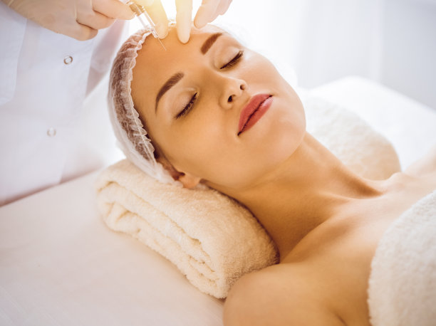 在阳光明媚的医疗中心，美女闭着眼睛接受美容注射。美容师医生用注射器给女性面部做美容手术。美容医学和外科的概念图片下载