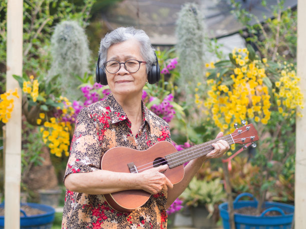 开朗的老年妇女，灰色短发，戴着眼镜，微笑着，站在花园里弹着尤克里里。图片下载