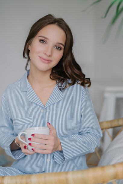 照片中漂亮的年轻女性直视镜头，穿着时尚的衣服，端着白色的杯子，里面装着芳香的饮料，在家里室内摆姿势，好好休息，和丈夫愉快地交谈。图片下载
