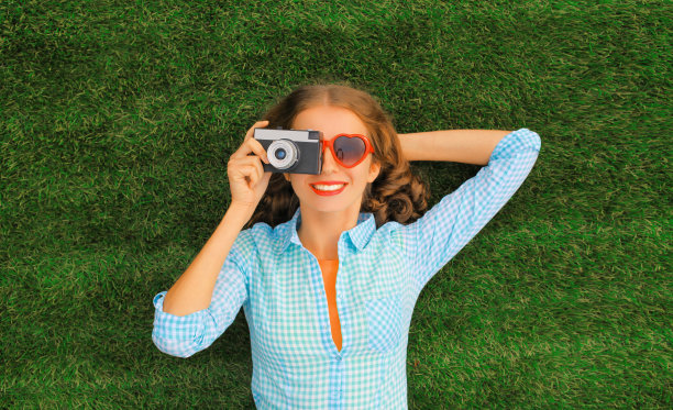 夏日肖像的快乐微笑的年轻女子摄影师拍照躺在公园的草地上图片下载