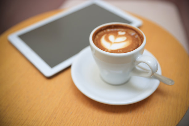 木桌上数码平板电脑旁的白色热咖啡杯。简单的工作空间或早上的咖啡休息时间/选择性专注图片下载