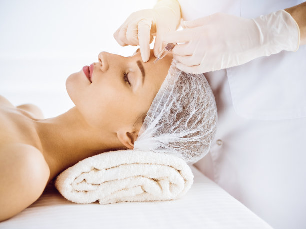 在阳光明媚的医疗中心，美女闭着眼睛接受美容注射。美容师医生用注射器给女性面部做美容手术。美容医学和外科的概念图片下载