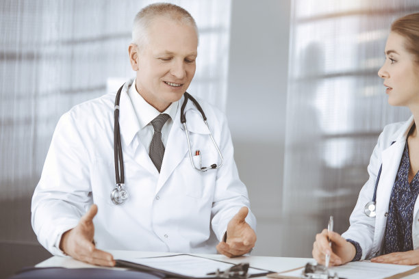 在阳光明媚的诊所里，老年医生和他的女同事坐在工作地点讨论目前的疾病治疗。医学中的团队合作图片下载