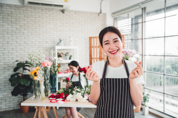 女花商坐在花店里，为一张宣传照片做着小心形手势。图片下载