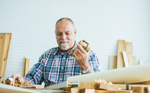 年长的老胡子和蔼的白种人或木匠，穿着格子衬衫，给孩子们DIY木制玩具，幸福地微笑着，独自坐在室内的家里。退休，爱好概念。图片下载