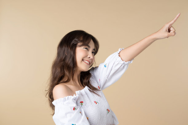 年轻的微笑的亚洲女人指着侧面的棕色背景复制空间。图片下载