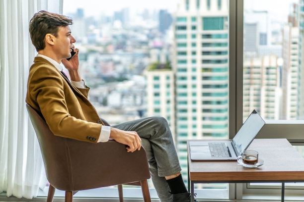 年轻的白人商人专业人士一边用笔记本电脑打电话，一边咨询一些文件，坐在办公桌前，一边用个人电脑工作，一边用智能手机通话图片下载