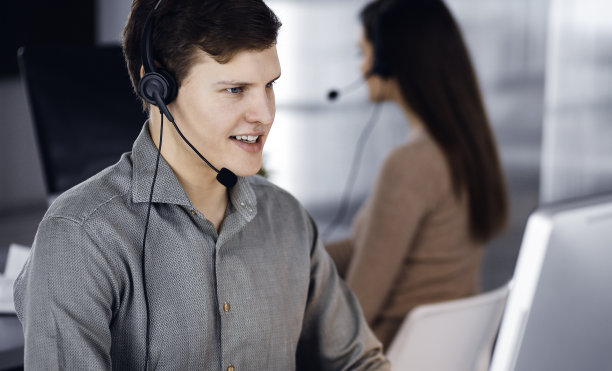 在一间现代化的办公室里，一个穿着绿色衬衫、戴着耳机的黑发年轻人正坐在办公桌前与一位女同事一起工作，与客户交谈。呼叫中心接线员正在工作图片下载